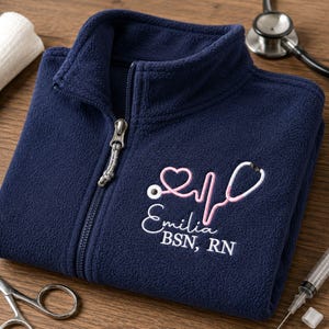 May include: A navy blue fleece jacket with a full-length zip and a high collar. Embroidered on the chest is a pink stethoscope forming a heart shape and an EKG line, with the white text "Emilia" and "BSN, RN" positioned underneath.