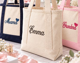 Könnte beinhalten: Drei Tragetaschen aus Segeltuch mit zwei Henkeln. Die linke Tasche ist cremefarben mit marineblauem Rand und dem gestickten Text "Mama" mit einem blauen Herz. Die mittlere Tasche ist cremefarben mit dem gestickten Namen "Emma". Die rechte Tasche ist cremefarben mit rosa Rand und dem gestickten Text "Grandma" mit einem rosa Herz.