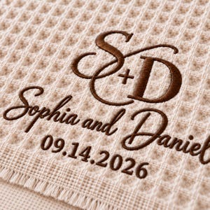 Parchment waffle throw featuring a personalized wedding embroidery positioned near the lower corner.
The design includes a large intertwined “S + D” monogram, “Sophia and Daniel” in script, and the date “09.14.2026” below.
All details are embroidered in deep brown thread for a clear contrast against the light Parchment fabric.
The elegant corner placement creates a personalized monogrammed wedding throw design.