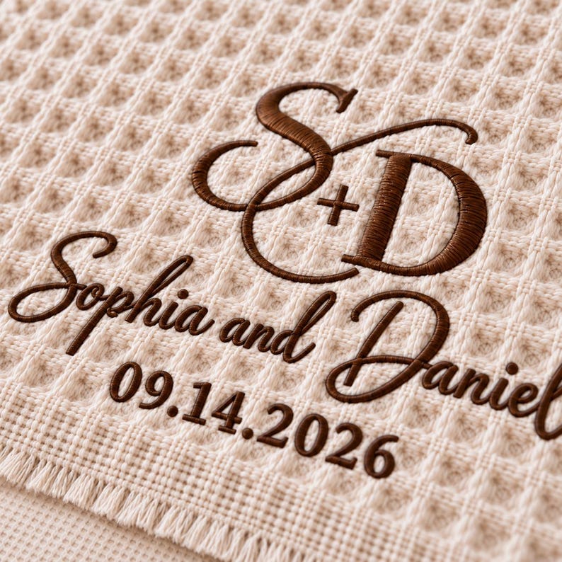 Parchment waffle throw featuring a personalized wedding embroidery positioned near the lower corner.
The design includes a large intertwined “S + D” monogram, “Sophia and Daniel” in script, and the date “09.14.2026” below.
All details are embroidered in deep brown thread for a clear contrast against the light Parchment fabric.
The elegant corner placement creates a personalized monogrammed wedding throw design.