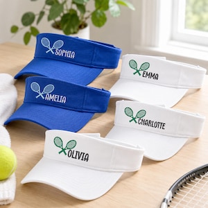 Könnte beinhalten: Eine Sammlung von fünf Sportvisieren in Königsblau und Weiß. Jedes Visier zeigt eine Stickerei von zwei gekreuzten Tennisschlägern und einen personalisierten Namen in schwarzem oder weißem Text, darunter Sophia, Amelia, Emma, Charlotte und Olivia.
