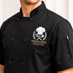 May include: A black double-breasted chef coat with a mandarin collar and short sleeves. The chest features a white embroidered logo of a chef hat, whisk, and spoon, with the gold text YOUR LOGO and CUSTOM TEXT printed below the graphic.