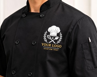Könnte beinhalten: Eine schwarze kurzärmelige Kochjacke mit doppelreihiger Vorderseite und schwarzen Knöpfen. Ein weißes gesticktes Logo auf der Brust zeigt eine Kochmütze, einen Schneebesen und einen Löffel, mit dem Text YOUR LOGO und CUSTOM TEXT darunter.