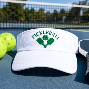 May include: A white visor featuring an embroidered green design on the front panel. The embroidery includes the word PICKLEBALL in an arched font above two crossed pickleball paddles and a single pickleball with circular perforations.