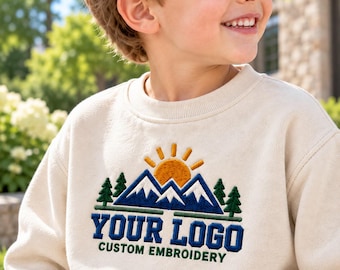Könnte beinhalten: Ein cremefarbenes Sweatshirt mit Rundhalsausschnitt und einer Stickerei, die eine gelbe Sonne über dunkelblauen Berggipfeln und grünen Tannen zeigt. Darunter befindet sich der blaue Schriftzug "YOUR LOGO" und der grüne Text "CUSTOM EMBROIDERY".
