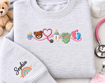 Könnte beinhalten: Ein hellgraues Sweatshirt mit Rundhalsausschnitt und gestickten Baby-Motiven, darunter ein Schnuller, ein Teddybär, ein rosa Stethoskop, ein Babyfläschchen, ein grüner Body mit Regenbogen und Babyfüßchen. Der Ärmel ist mit dem Namen Sophia bestickt.
