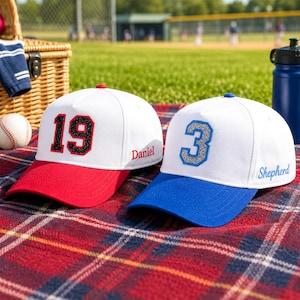 May include: Two white baseball caps with colored brims and glittery numbers on the front. One cap has a red brim and the number 19 with the name Daniel embroidered on the side. The other cap has a blue brim and the number 3 with the name Shepherd embroidered on the side.
