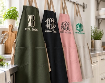 Könnte beinhalten: Vier Schürzen hängen an einer Metallstange, jede mit zwei Vordertaschen und beigen Nackenbändern. Die olivgrüne Schürze zeigt SCB EST. 2026, die schwarze IGC Custom Text, die rosa Schürze SBL MRS. CARTER und die beige EME THE MILLER KITCHEN.