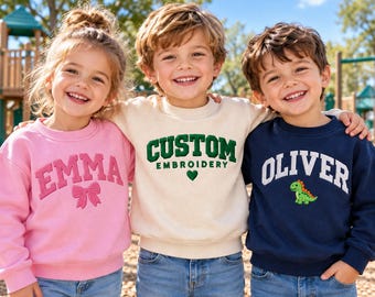 Könnte beinhalten: Drei Kinder tragen Rundhals-Sweatshirts. Das linke ist rosa mit dem Namen EMMA und einer Schleife. Das mittlere ist cremefarben mit dem Text CUSTOM EMBROIDERY und einem Herz. Das rechte ist marineblau mit dem Namen OLIVER und einem Dinosaurier.