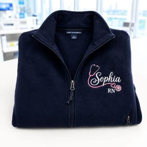 May include: A navy blue fleece jacket with a full-length front zip and a high collar. The chest features embroidered text that reads Sophia in white script, accompanied by a pink stethoscope graphic and the letters RN in white block font.