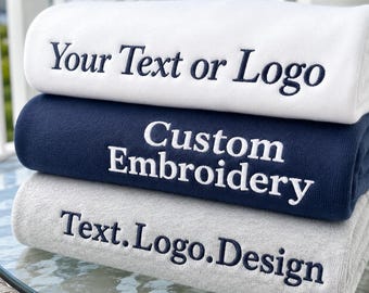 Puede incluir: Una pila de tres mantas dobladas. La manta blanca superior presenta un texto bordado en azul oscuro que dice Your Text or Logo. La manta azul marino central tiene un texto bordado en blanco que dice Custom Embroidery. La manta gris inferior tiene un texto bordado en azul oscuro que dice Text.Logo.Design.