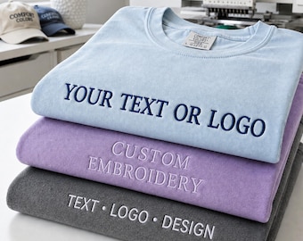 Könnte beinhalten: Ein Stapel aus drei Sweatshirts mit Rundhalsausschnitt in Hellblau, Lavendel und Dunkelgrau. Das hellblaue Sweatshirt zeigt die dunkelblaue Stickerei YOUR TEXT OR LOGO. Das lavendelfarbene trägt CUSTOM EMBROIDERY und das graue TEXT • LOGO • DESIGN.