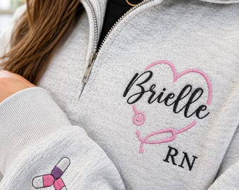 Puede incluir: Una sudadera gris claro con cremallera corta que presenta un diseño bordado de un estetoscopio en forma de corazón rosa con el nombre Brielle y las letras RN en negro. La manga muestra un gráfico bordado de tres pastillas en rosa, blanco y morado.
