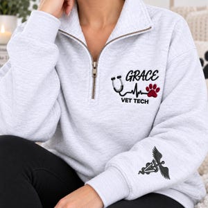 Puede incluir: Una sudadera gris claro con cremallera corta y un diseño bordado en el pecho con un estetoscopio, una línea de latido, una huella roja y el texto GRACE VET TECH. La manga izquierda presenta un símbolo de caduceo negro bordado.