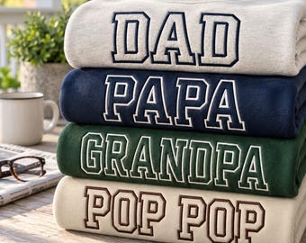 Könnte beinhalten: Ein Stapel aus vier gefalteten Rundhals-Sweatshirts in Hellgrau, Marineblau, Dunkelgrün und Creme. Jedes Sweatshirt ist auf der Vorderseite mit gestickten Blockbuchstaben versehen, die DAD, PAPA, GRANDPA und POP POP lauten.