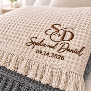 Parchment and Gusty Grey waffle throws featuring a personalized wedding monogram embroidered near the lower corner.
The Parchment throw displays an elegant “S + D” monogram with “Sophia and Daniel” and the date “09.14.2026” stitched below.
Rich brown thread creates the initials, script names, and date with a clean raised embroidery finish.
The corner placement keeps the personalized design visible while highlighting the textured waffle fabric and fringe.