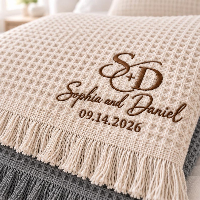 Parchment and Gusty Grey waffle throws featuring a personalized wedding monogram embroidered near the lower corner.
The Parchment throw displays an elegant “S + D” monogram with “Sophia and Daniel” and the date “09.14.2026” stitched below.
Rich brown thread creates the initials, script names, and date with a clean raised embroidery finish.
The corner placement keeps the personalized design visible while highlighting the textured waffle fabric and fringe.