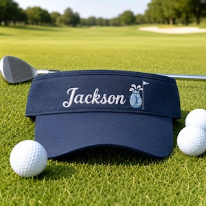 May include: A navy blue golf visor featuring the embroidered name Jackson in white script lettering, accompanied by a small light blue golf bag icon with clubs and a flag. The visor is positioned on a green golf course with white golf balls.
