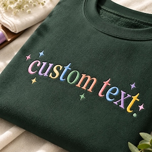 Puede incluir: Una sudadera de cuello redondo en color verde bosque oscuro con el texto bordado "custom text" en una fuente redondeada multicolor. El diseño está acentuado con pequeñas estrellas bordadas de colores dispersas por las letras.