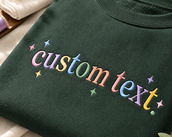 Könnte beinhalten: Ein dunkelwaldgrüner Rundhals-Pullover mit den gestickten Wörtern custom text in einer bunten Schriftart. Das Design wird durch kleine, gestickte vierzackige Sterne in verschiedenen Pastellfarben akzentuiert, die um den Text herum verteilt sind.