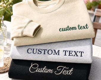 Könnte beinhalten: Ein Stapel aus drei Sweatshirts mit Rundhalsausschnitt in Beige, Hellgrau und Schwarz. Das beige Sweatshirt hat einen grünen Stickschriftzug mit dem Text custom text. Das graue Sweatshirt zeigt dunkelblauen Sticktext mit CUSTOM TEXT. Das schwarze Sweatshirt trägt cremefarbenen Sticktext mit Custom Text.