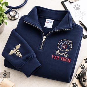 May include: A navy blue quarter-zip fleece pullover featuring an embroidered logo with a cat, dog, heart, and stethoscope. The text reads Emily VET TECH. The sleeve displays a gold caduceus symbol with a paw print. A Jerzees NuBlend tag is visible.