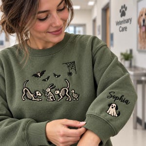 Puede incluir: Una sudadera de cuello redondo color verde oliva con un diseño bordado de esqueletos de animales, incluyendo un perro, un conejo, un gato y un pato, con murciélagos y una telaraña. La manga tiene bordado el nombre Sophia y un perro fantasma.