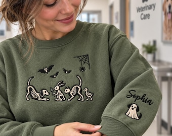 Könnte beinhalten: Ein olivgrünes Sweatshirt mit Rundhalsausschnitt und einer Stickerei von Tierskeletten, darunter ein Hund, ein Hase, eine Katze und eine Ente, sowie Fledermäusen und einem Spinnennetz. Der Ärmel ist mit dem Namen Sophia und einem Geisterhund bestickt.