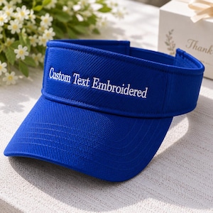 May include: A royal blue sun visor featuring the white embroidered text Custom Text Embroidered on the front panel. The visor has a structured brim and an adjustable strap at the back, constructed from a durable, textured fabric material.