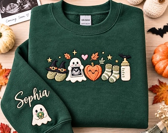 Könnte beinhalten: Ein dunkelgrüner Rundhals-Pullover mit gestickten Baby-Motiven zum Thema Halloween, darunter Geister, ein Kürbis, Babysocken und ein Fläschchen. Auf dem Ärmel ist der Name Sophia und ein kleiner Geist mit Schnuller eingestickt.