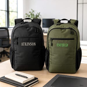 May include: Two backpacks, one black and one olive green, each featuring a front zippered pocket with embroidered text. The black backpack displays the name ATKINSON in white, while the olive green backpack features the name ENFIELD in green.
