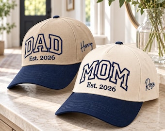 Könnte beinhalten: Zwei beige Baseballkappen mit marineblauem Schirm. Eine Kappe zeigt den marineblauen Schriftzug DAD Est. 2026 und den Namen Henry an der Seite. Die andere Kappe zeigt den marineblauen Schriftzug MOM Est. 2026 und den Namen Rose an der Seite.