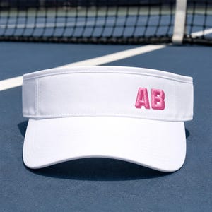 May include: A white visor featuring the letters AB embroidered in pink on the front panel. The visor has a curved brim with visible stitching and an adjustable strap at the back, designed for sun protection during outdoor athletic activities.