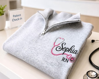 Puede incluir: Una sudadera gris claro con cremallera de un cuarto y un diseño bordado en el pecho. El bordado incluye un estetoscopio rosa que forma un corazón, el nombre Sophia en letra cursiva negra y las letras RN en fuente negra debajo del nombre.