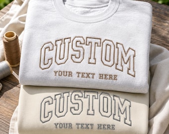 Könnte beinhalten: Zwei Rundhals-Sweatshirts, eines hellgrau und eines beige, mit gesticktem Text. Das obere Sweatshirt zeigt das Wort CUSTOM in braunen Blockbuchstaben mit dem Text YOUR TEXT HERE darunter. Das untere Sweatshirt zeigt denselben Text in Grau.