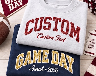 Könnte beinhalten: Ein hellgraues Sweatshirt mit Rundhalsausschnitt und dem roten Glitzertext CUSTOM und Custom Text, das über einem marineblauen Sweatshirt mit goldenem Glitzertext GAME DAY und dem weißen Text Sarah 2026 in College-Schriftart liegt.