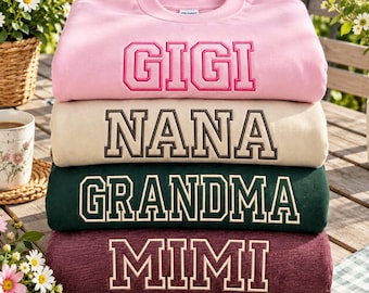 Könnte beinhalten: Ein Stapel aus vier Sweatshirts mit Rundhalsausschnitt in Rosa, Beige, Dunkelgrün und Kastanienbraun. Jedes Sweatshirt ist auf der Brust mit gestickten Blockbuchstaben versehen, die GIGI, NANA, GRANDMA und MIMI in Kontrastfarben zeigen.