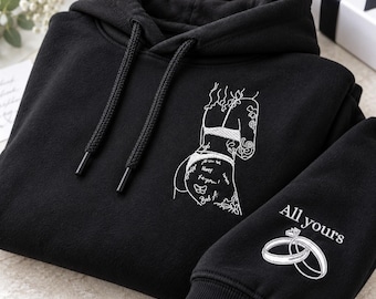 Könnte beinhalten: Ein schwarzer Kapuzenpullover mit weißer Linienstickerei einer Figur in Unterwäsche auf der Brust. Der Ärmel zeigt den Text All yours über einer gestickten Grafik aus zwei ineinandergreifenden Ringen mit Diamantbesatz.