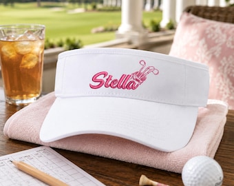 Könnte beinhalten: Ein weißes Golf-Visier mit dem in pinker Schreibschrift gestickten Namen Stella, begleitet von einer kleinen pinken Grafik einer Golftasche mit Schlägern. Das Visier besteht aus einfarbigem weißem Stoff mit gebogenem Schirm und verstellbarem Riemen.