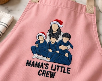 Könnte beinhalten: Eine hellrosa Schürze mit beigen Trägern und einer Stickerei, die eine Person mit Weihnachtsmütze und drei Kinder in blauer Kleidung zeigt. Unter den Figuren ist der Text MAMA'S LITTLE CREW in schwarzem Garn eingestickt.