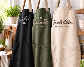 Könnte beinhalten: Drei Schürzen in Schwarz, Olivgrün und Beige, jeweils mit zwei Vordertaschen und beigen Nackenbändern. Die schwarze Schürze trägt die Aufschrift Sophia CHEF, die grüne CHEF Custom Text EST. 2026 und die beige The Smith Kitchen MADE WITH LOVE.