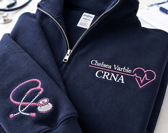 Könnte beinhalten: Ein marineblauer Fleece-Pullover mit Viertelreißverschluss und weiß-rosa Stickerei. Auf der Brust befindet sich der Text Chelsea Varble CRNA mit einem rosa Herz und einer EKG-Linie. Der Ärmel zeigt ein gesticktes Stethoskop-Motiv in Rosa und Weiß.