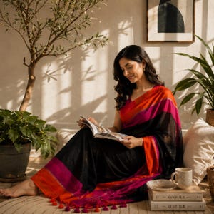 May include: A black saree featuring vibrant orange and magenta horizontal stripes along the border and pallu, finished with decorative tassels. The fabric has a subtle texture, and the garment is draped in a traditional style with a matching blouse.