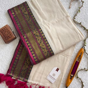Peut inclure: Un textile blanc cassé doté d'une large bordure à motifs floraux dorés et magenta. Le bord est fini avec des pompons magenta. Une étiquette blanche avec le texte Sutisancha est attachée. Une navette en bois et une petite boîte sont présentes.
