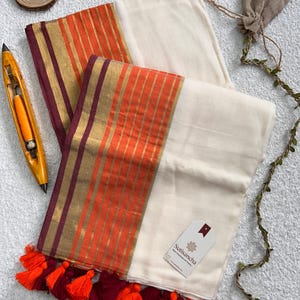 May include: An off-white textile featuring a wide border with alternating orange and gold vertical stripes, finished with a deep maroon edge. The bottom hem is adorned with a row of alternating bright orange and deep maroon fabric tassels.