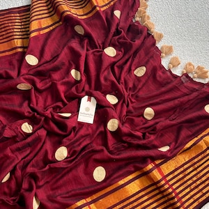 May include: A deep maroon fabric featuring a pattern of gold circular dots, finished with a wide border of horizontal gold and maroon stripes. The edge is adorned with beige tassels, and a white tag labeled Sutisancha is attached to the center.