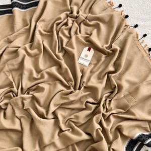 May include: A beige fabric textile featuring a black and white patterned border along the edges. The fabric is draped with folds and includes small black and beige tassels along one side. A white tag with the text Sutisancha is attached.