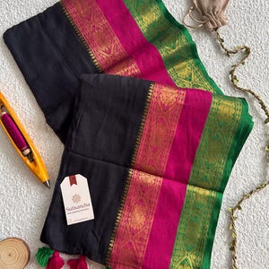 Peut inclure: Un textile en tissu noir doté d'une large bordure magenta et d'un liseré vert, tous deux ornés de motifs métalliques dorés complexes. Le tissu présente de petits pompons verts et magenta sur le bord inférieur et une étiquette blanche Sutisancha.