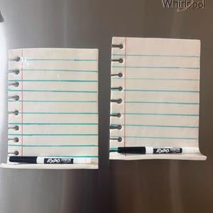 May include: Two white ceramic dry-erase boards shaped like notebook paper with blue horizontal lines and a red vertical margin line. Each board features a row of circular cutouts along the left side and a black Expo dry-erase marker resting on a ledge.