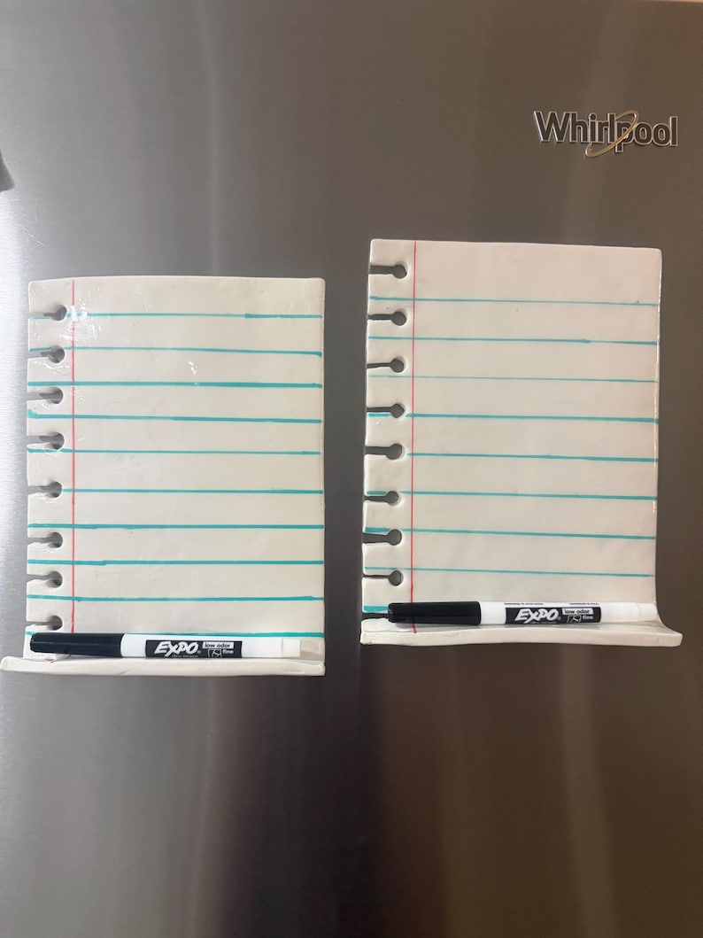 May include: Two white ceramic dry-erase boards shaped like notebook paper with blue horizontal lines and a red vertical margin line. Each board features a row of circular cutouts along the left side and a black Expo dry-erase marker resting on a ledge.